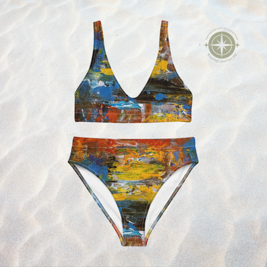 A two-piece bikini set with an abstract, textured paint-stroke pattern in multiple colors, laid flat on a textured white sand background. A compass logo is visible in the top right corner.
