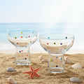 Two glass tumblers with colorful polka dots on a sandy beach with shells and a starfish.