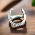 A handmade silver ring in the shape of a horseshoe, with the name 'Rebecca' stamped on it, displayed on a wooden surface.