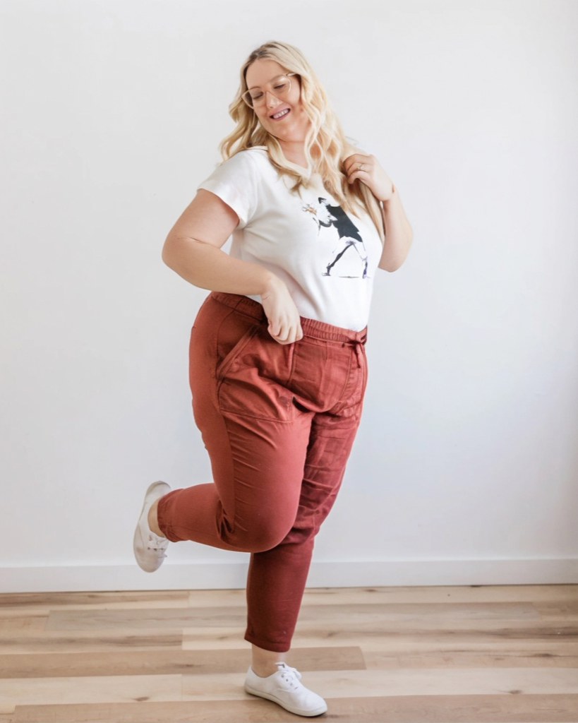 Woman wearing a white t-shirt with a graphic design and red pants, standing on a wooden floor against a white wall.