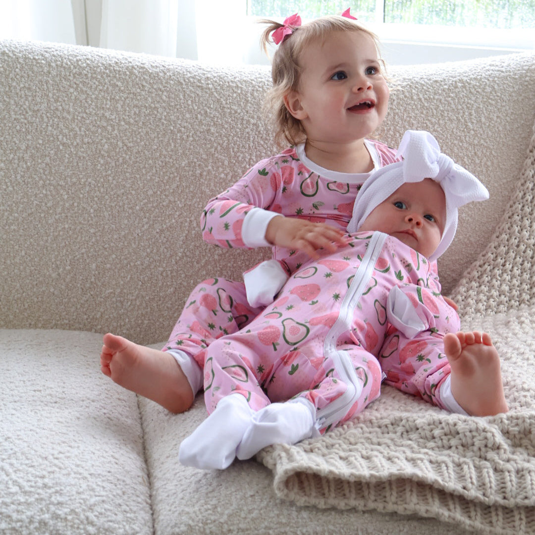 Two children in pink pajamas sitting on a couch with a window in the background.
