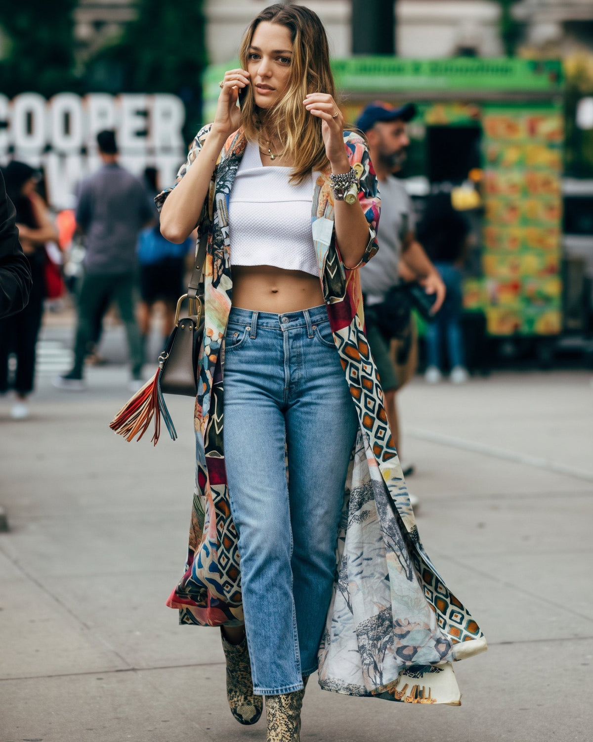 A woman walking on a city street wearing a white crop top, blue jeans, a long patterned bohemian duster cardigan, and leopard-print ankle boots.