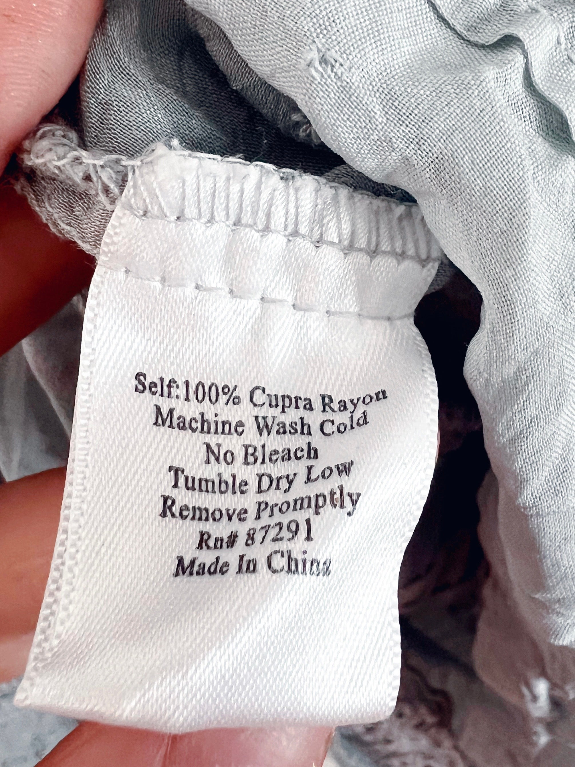 Close-up of a fabric garment label sewn onto a light gray textile, held by a hand. The label reads: 'Self: 100% Cupra Rayon Machine Wash Cold No Bleach Tumble Dry Low Remove Promptly Rs# 87291 Made In China'.