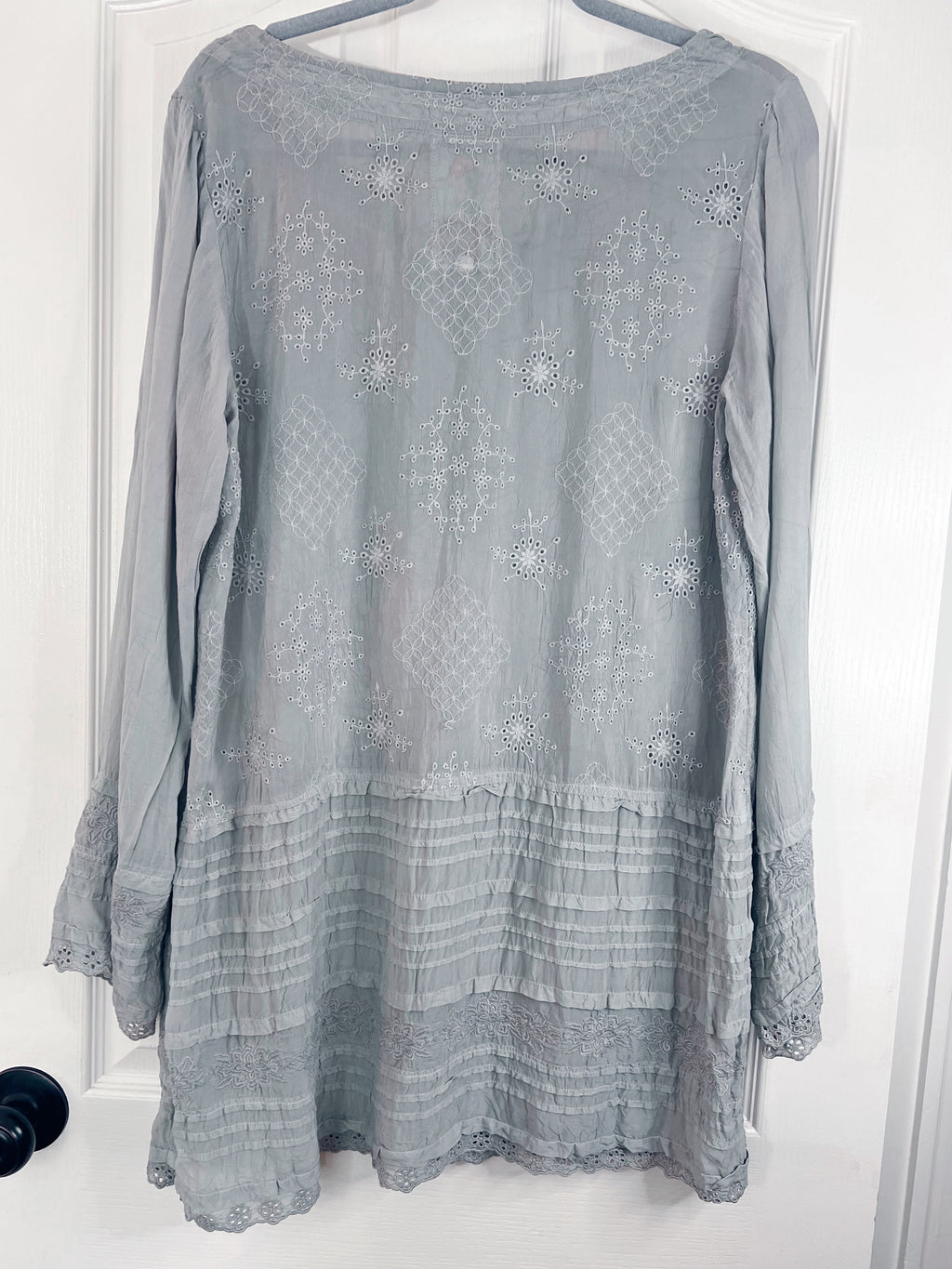 A light gray long-sleeve tunic top with intricate embroidered patterns and a tiered ruffled hem, hanging on a hanger against a white door.