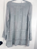 A light gray long-sleeve tunic top with intricate embroidered patterns and a tiered ruffled hem, hanging on a hanger against a white door.