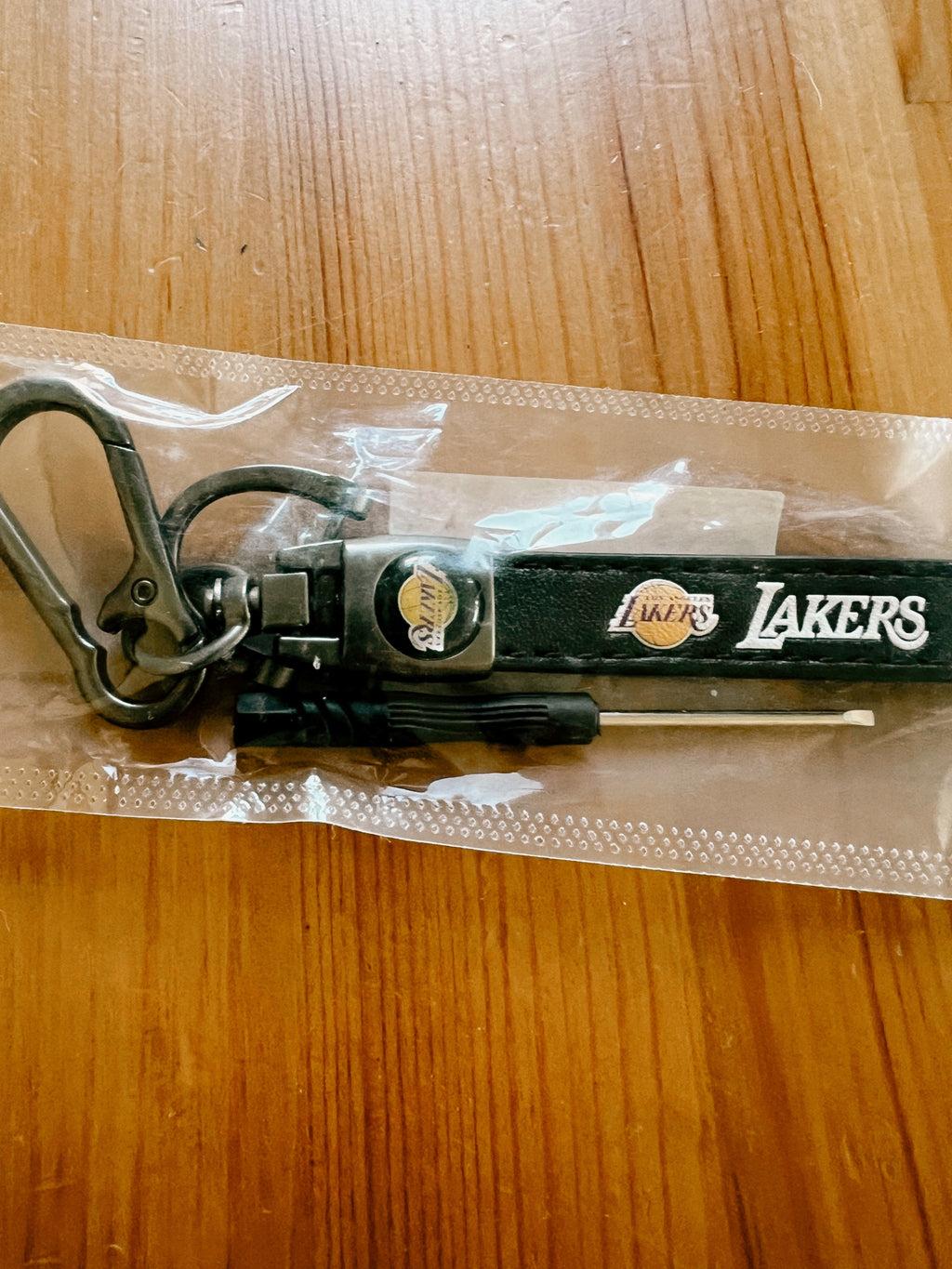 A black Los Angeles Lakers keychain with a carabiner and keyring, still sealed in its clear plastic packaging, placed on a wooden tabletop.