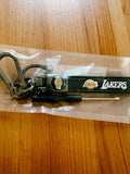 A black Los Angeles Lakers keychain with a carabiner and keyring, still sealed in its clear plastic packaging, placed on a wooden tabletop.