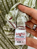 A close-up of a person's hand holding a folded green and white patterned garment, showing the sewn-in label that reads 'Lucky Brand DUNGAREES BY GENE MONTESANO STYLED IN U.S.