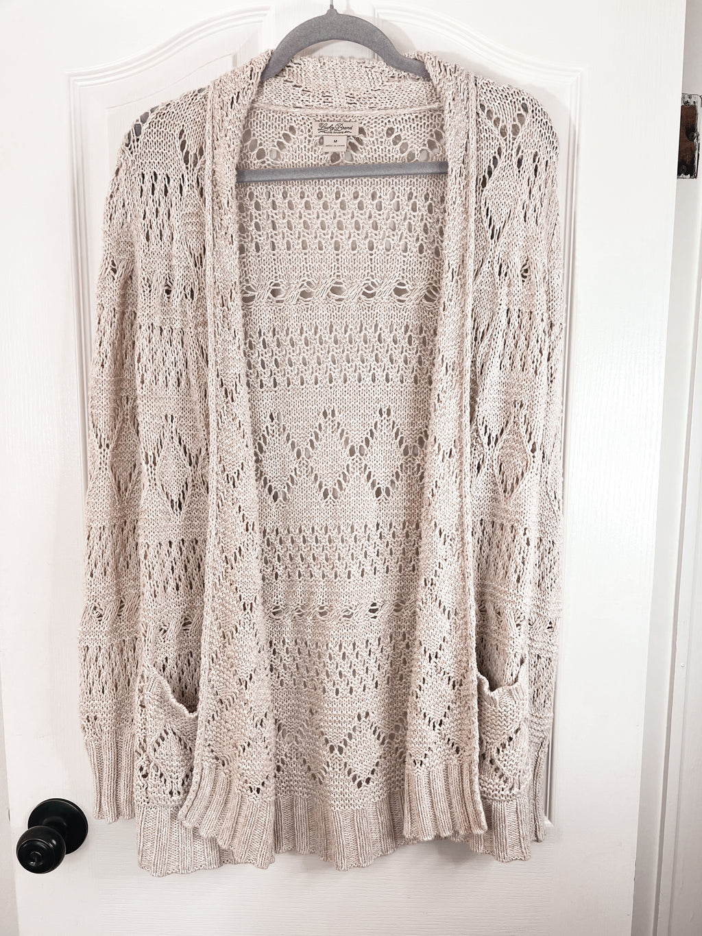 A light beige open-front knit cardigan sweater with an intricate openwork pattern, hanging on a hanger against a white door.