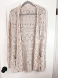 A light beige open-front knit cardigan sweater with an intricate openwork pattern, hanging on a hanger against a white door.