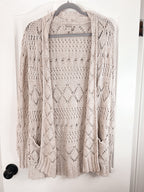 A light beige open-front knit cardigan sweater with an intricate openwork pattern, hanging on a hanger against a white door.
