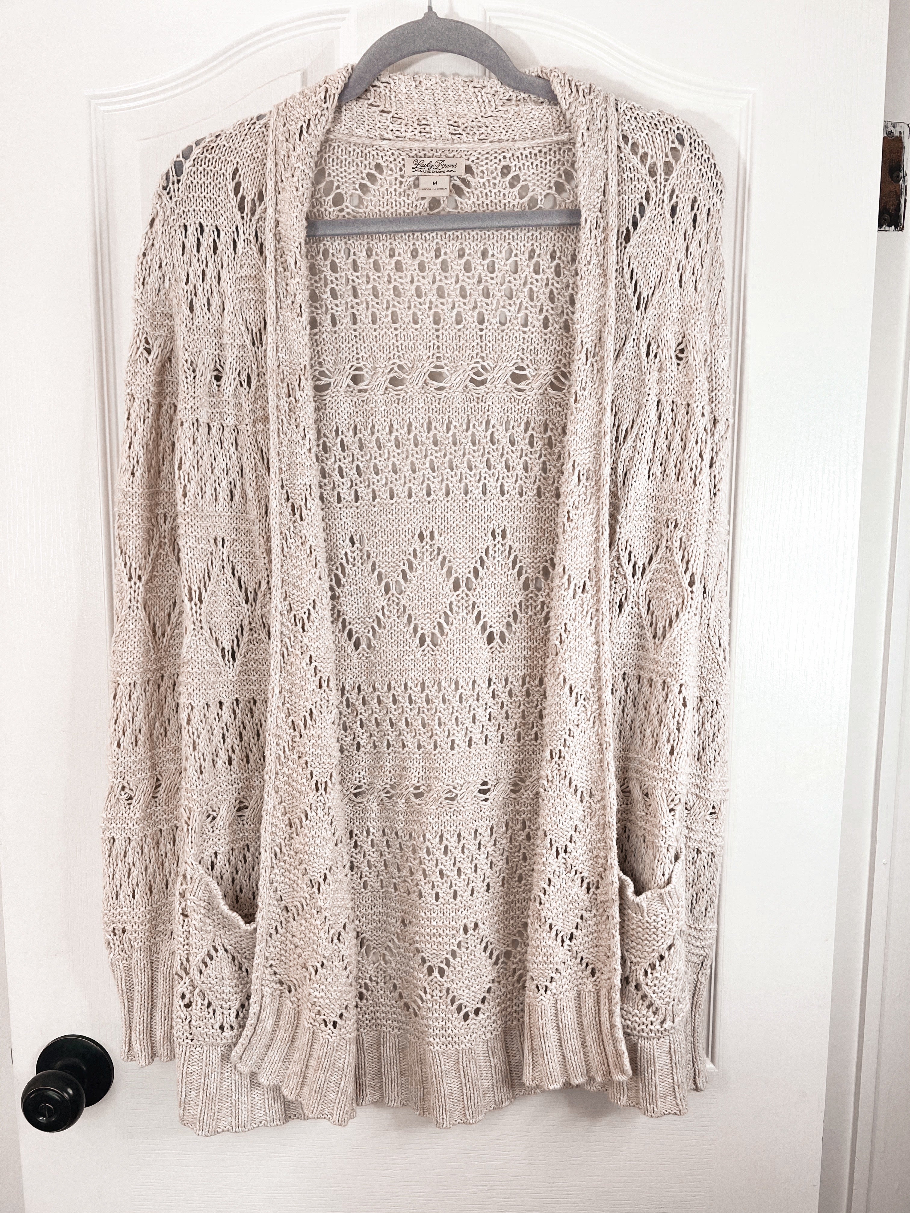 A light beige open-front knit cardigan sweater with an intricate openwork pattern, hanging on a hanger against a white door.