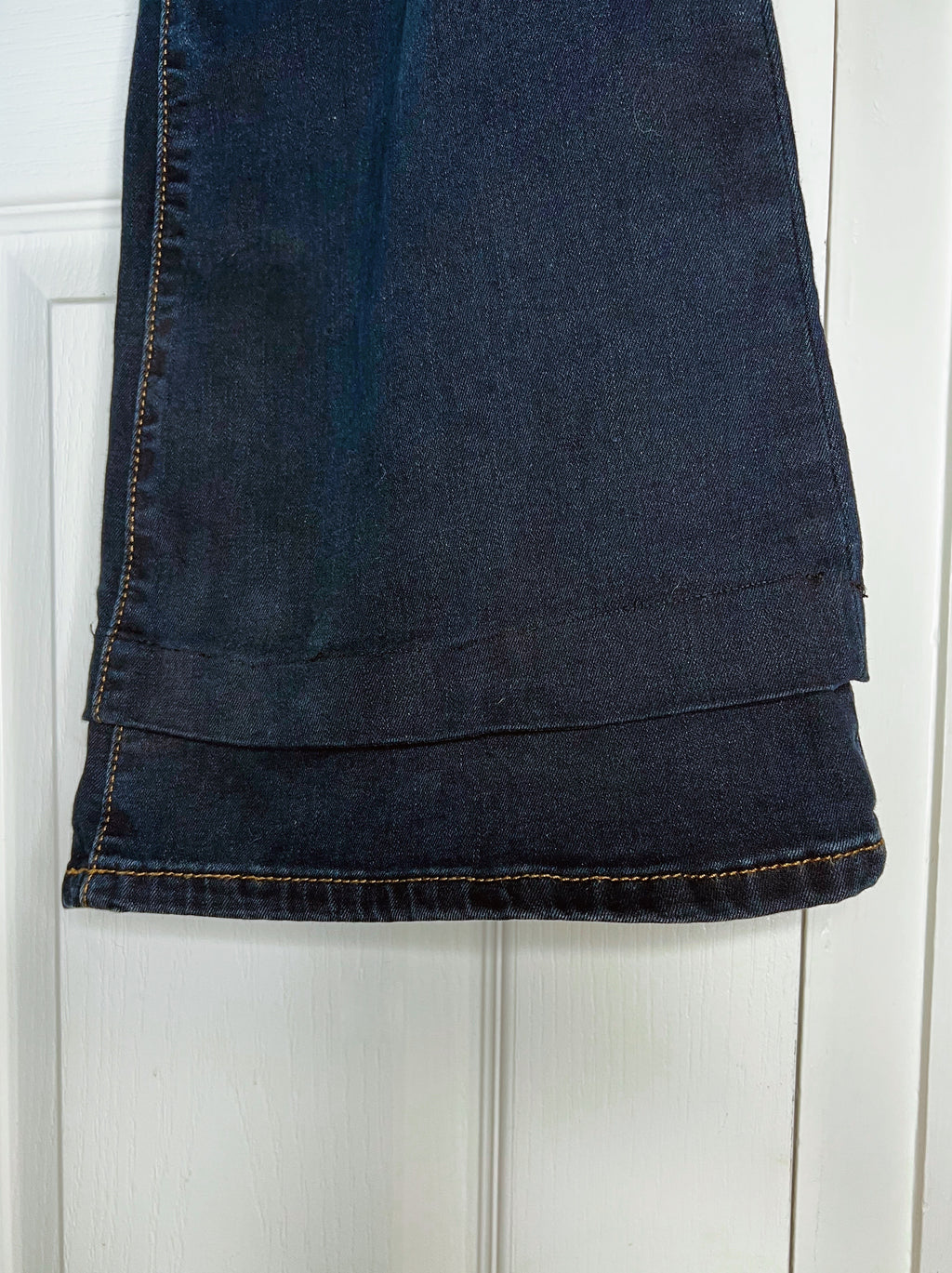A close-up of a dark blue denim garment, likely a skirt or pants leg, hanging against a white paneled door. The fabric shows dark blue wash details, visible stitching, and a folded hem.