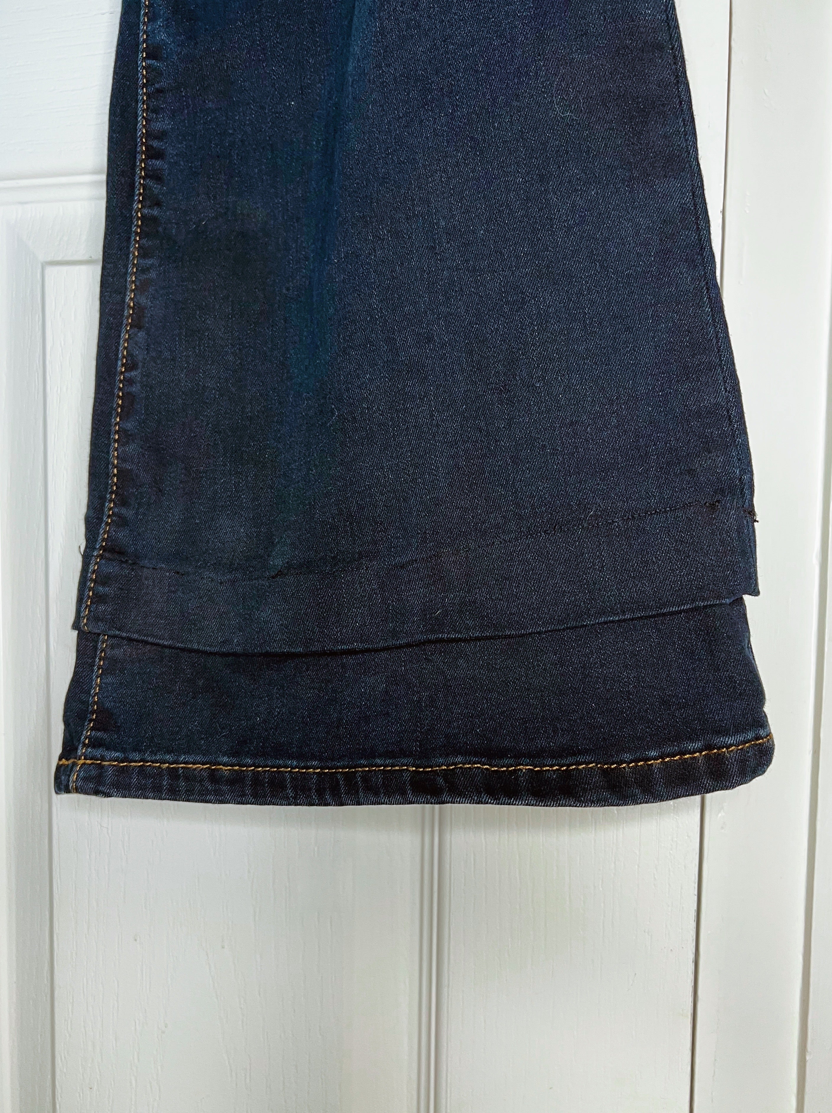 A close-up of a dark blue denim garment, likely a skirt or pants leg, hanging against a white paneled door. The fabric shows dark blue wash details, visible stitching, and a folded hem.