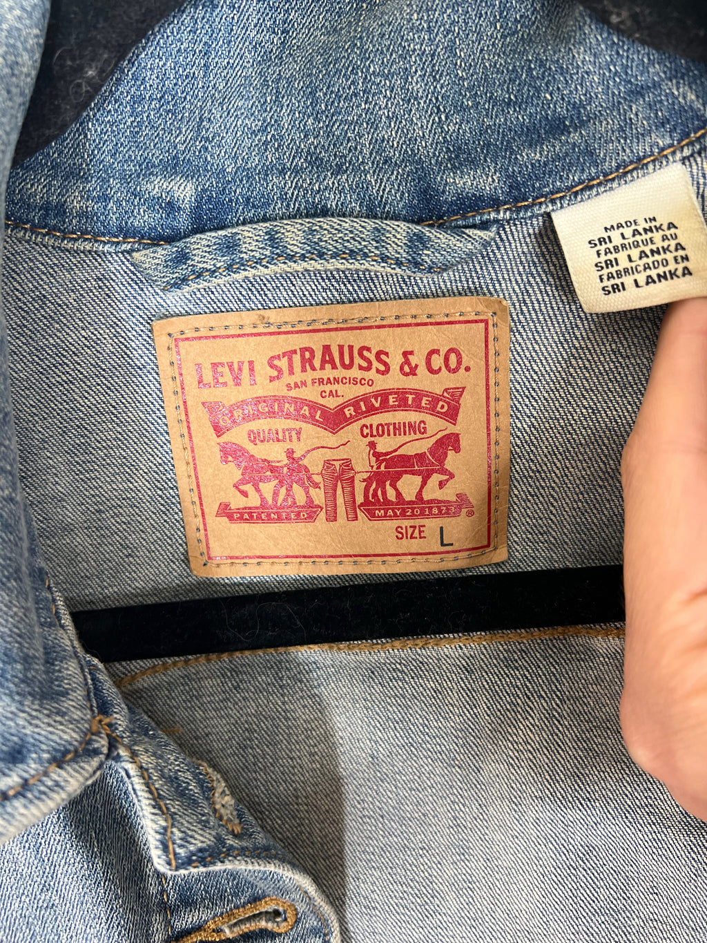 Close-up of a denim jacket's inner neck label showing the classic tan leather Levi's Strauss & Co. patch, with a hand holding a country-of-origin tag next to it.