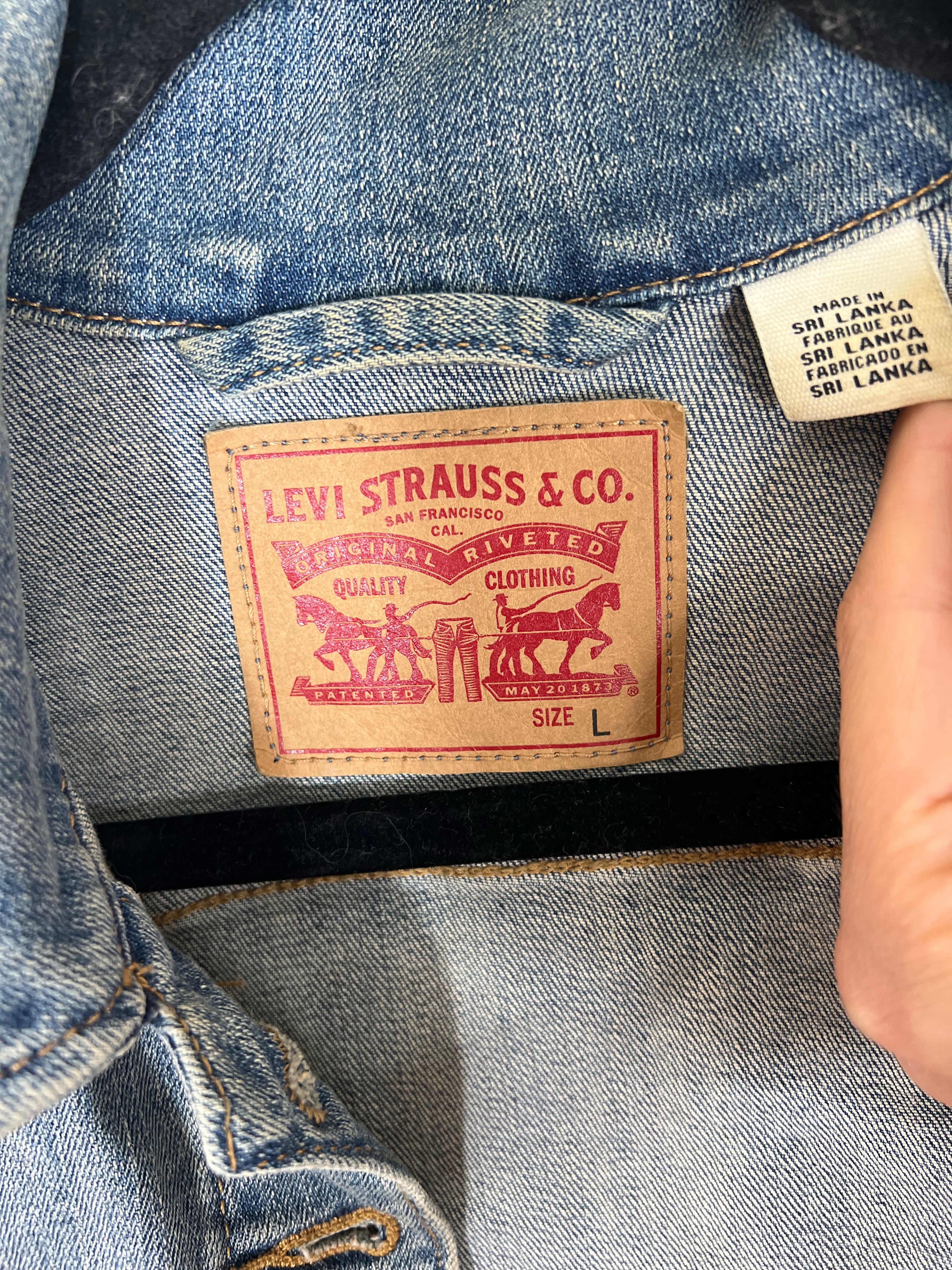 Close-up of a denim jacket's inner neck label showing the classic tan leather Levi's Strauss & Co. patch, with a hand holding a country-of-origin tag next to it.