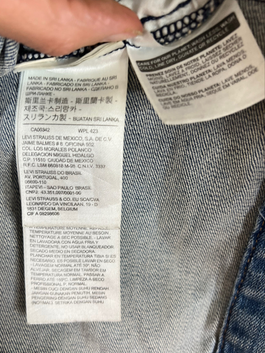 Close-up of a denim garment's interior label area with a finger pointing at the sewn-in care and size labels. The fabric is blue denim, and the labels are white with black text.