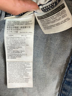 Close-up of a denim garment's interior label area with a finger pointing at the sewn-in care and size labels. The fabric is blue denim, and the labels are white with black text.