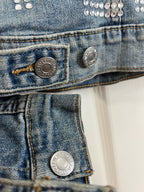 Close-up detail of a blue denim jacket showing silver metal buttons, contrast stitching, and rhinestone embellishments on the chest.