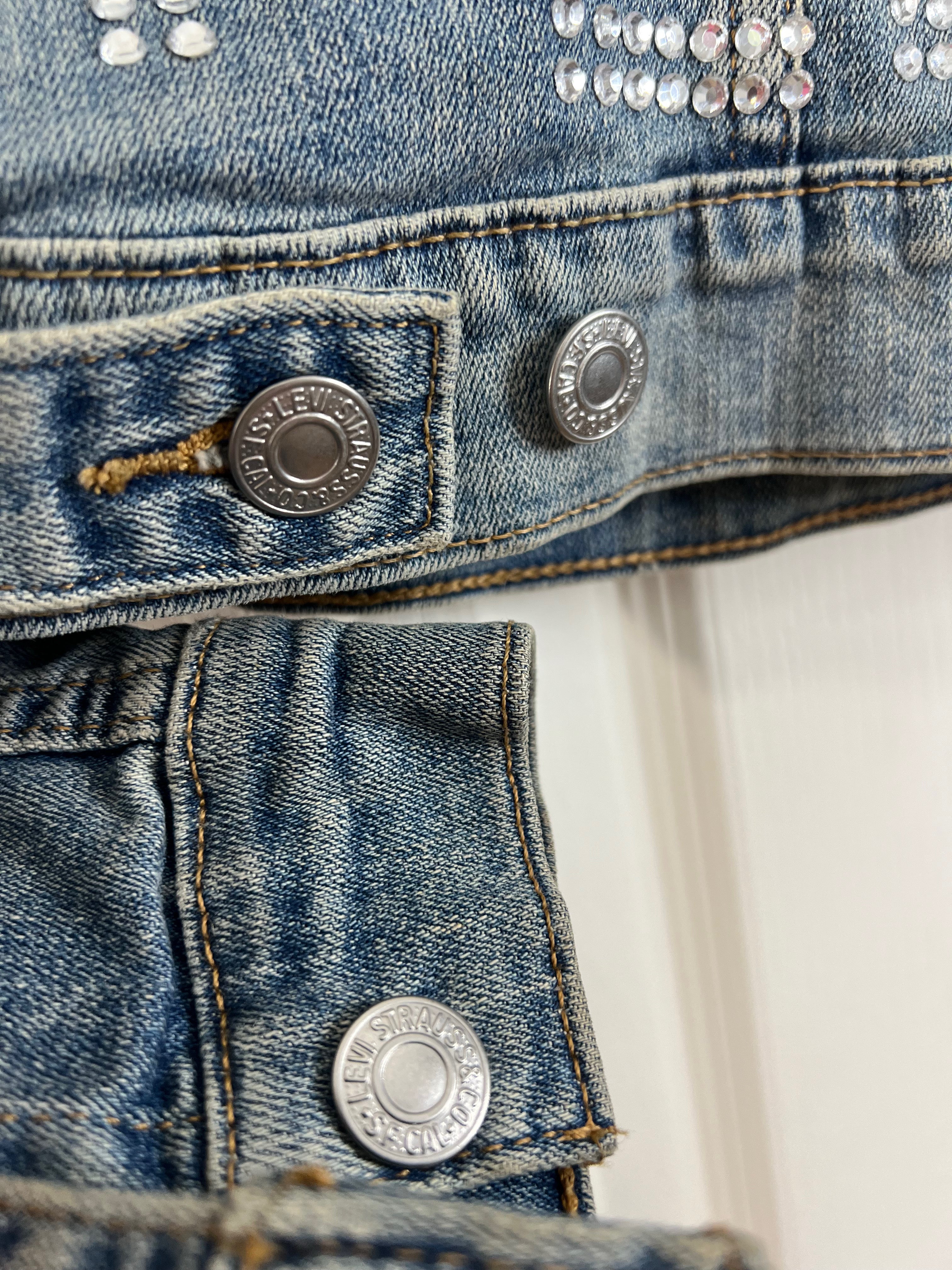 Close-up detail of a blue denim jacket showing silver metal buttons, contrast stitching, and rhinestone embellishments on the chest.