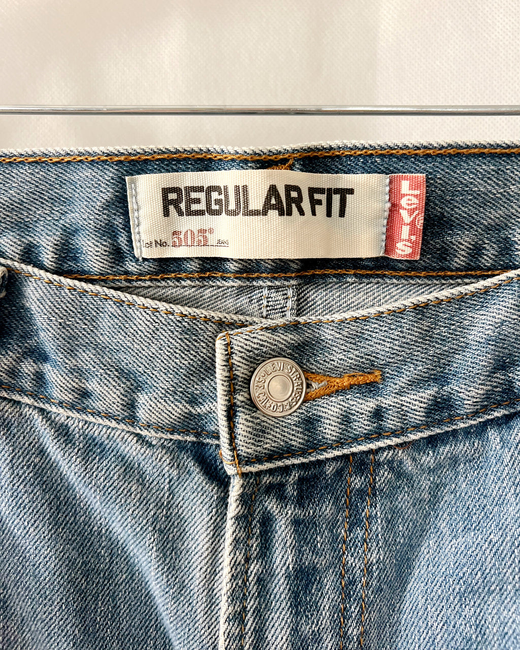 Close-up of the waistband of Levi's Regular Fit 505 jeans, showing the white and red brand labels, a silver metal button, and light blue denim fabric.