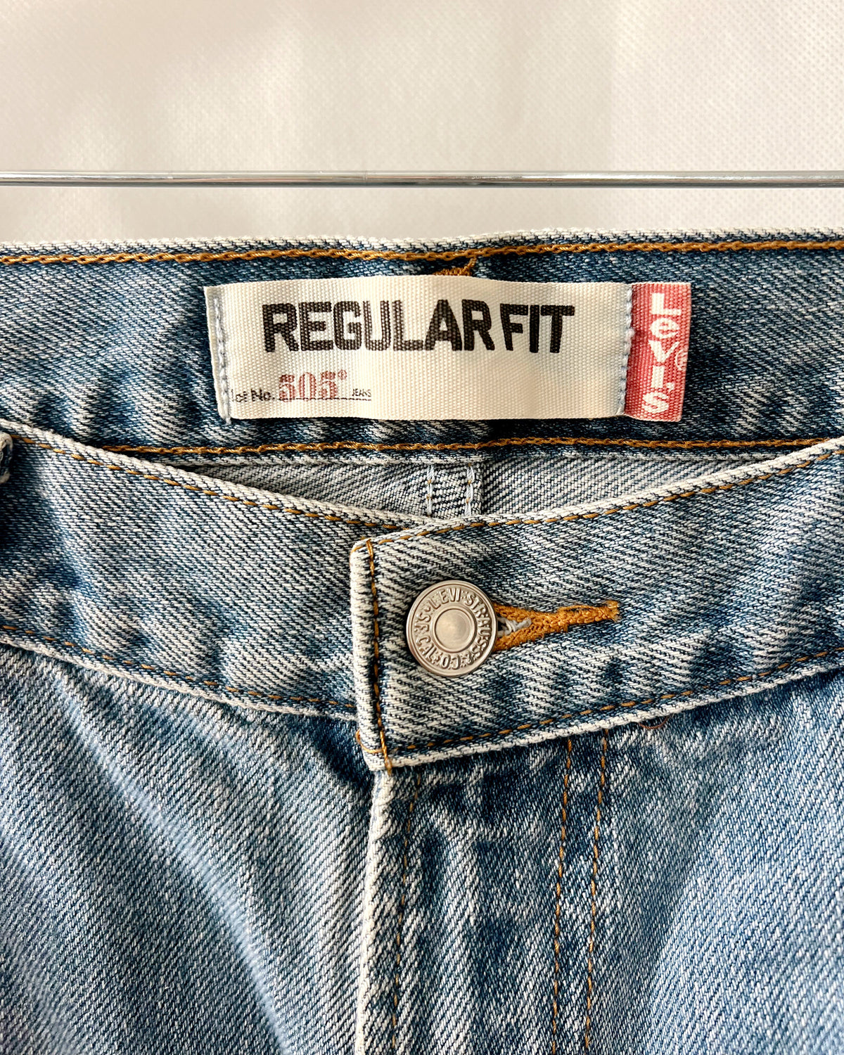 Close-up of the waistband of Levi's Regular Fit 505 jeans, showing the white and red brand labels, a silver metal button, and light blue denim fabric.