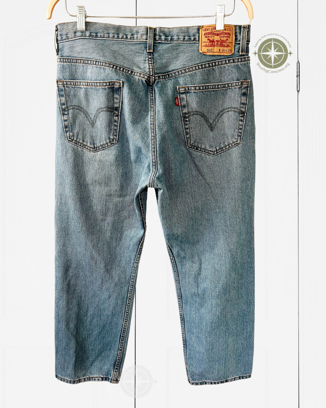 Back view of light blue Levi's denim jeans hanging on a wooden hanger against a white paneled background, showing the classic red tab and leather patch on the waistband.