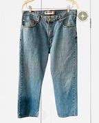 A pair of light blue denim jeans with a faded wash, hanging from a wooden hanger against a white paneled background. The brand name 'SICILIANI' is visible on the waistband label.