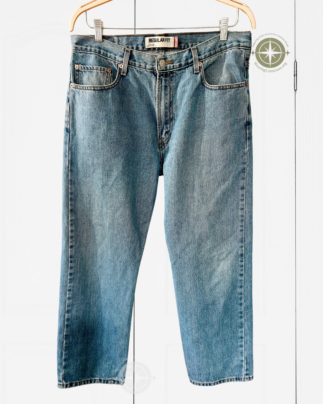 A pair of light blue denim jeans with a faded wash, hanging from a wooden hanger against a white paneled background. The brand name 'SICILIANI' is visible on the waistband label.