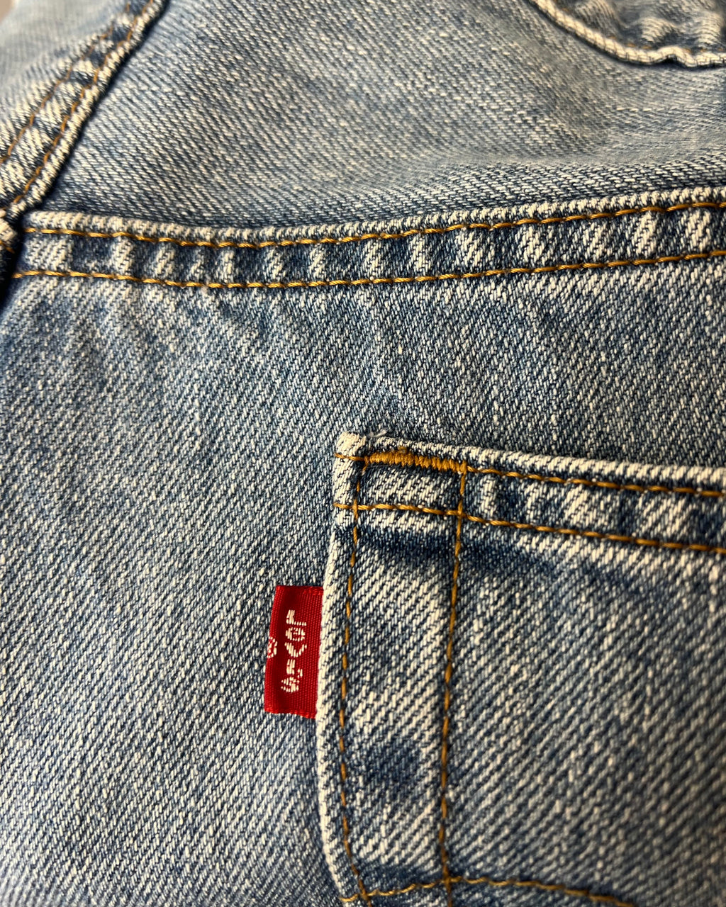 Close-up of a light blue denim garment, showing the back pocket with orange contrast stitching and a red Levi's waist tab.