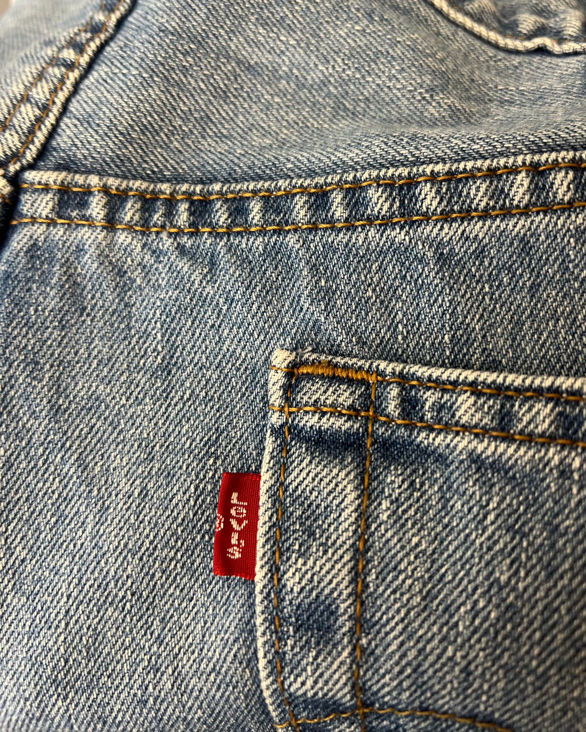 Close-up of a light blue denim garment, showing the back pocket with orange contrast stitching and a red Levi's waist tab.