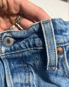 Close-up of a person's hand holding up the waistband of light blue denim jeans, showing the silver metal button, belt loops, and copper-toned rivets.