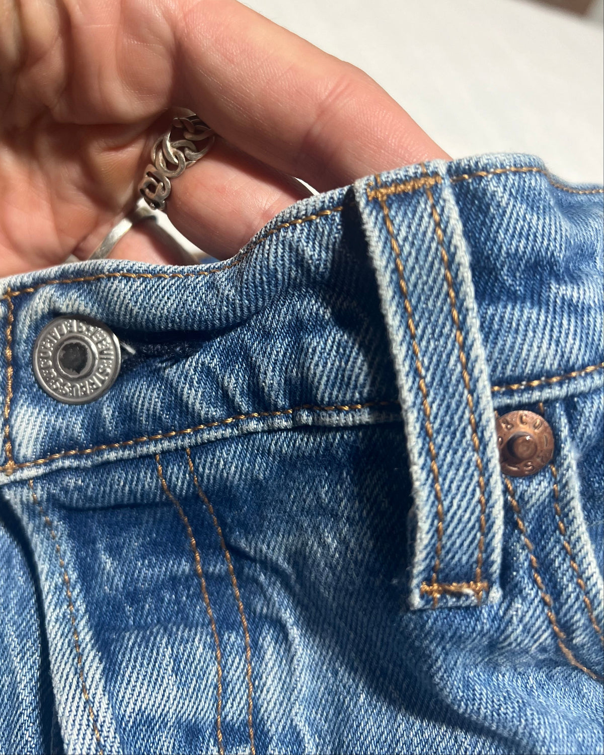 Close-up of a person's hand holding up the waistband of light blue denim jeans, showing the silver metal button, belt loops, and copper-toned rivets.