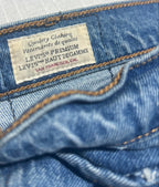 Close-up of a denim waistband showing the interior label for Levi's Premium, with a finger holding the fabric open. The blue denim and tan contrast stitching are clearly visible against a light gray background.