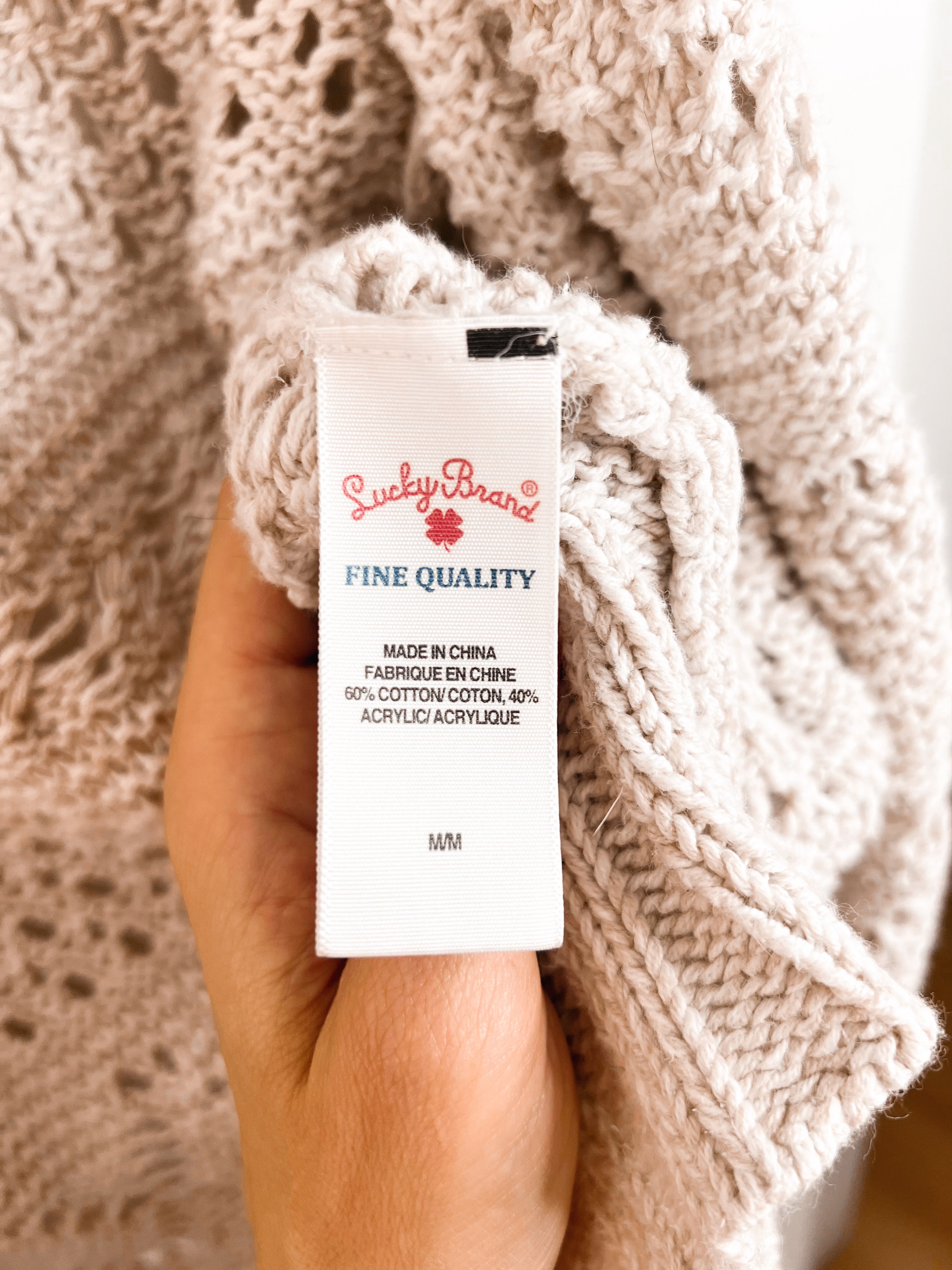 A hand holds a white fabric tag for a chunky knit sweater. The tag is branded 'Suckly Brand' with 'FINE QUALITY' written on it.