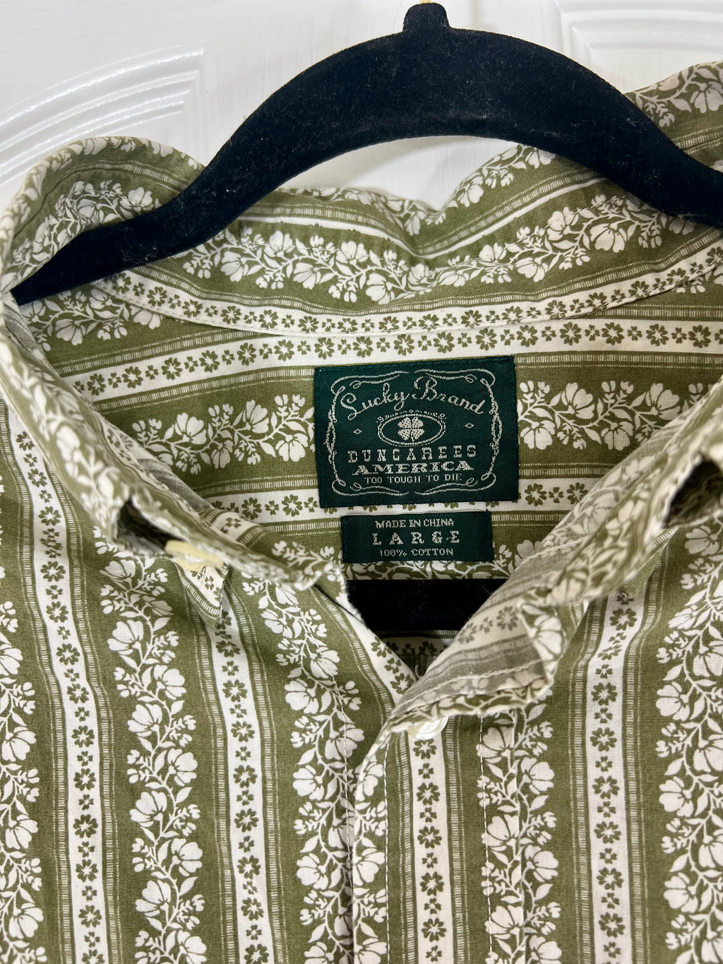 A close-up of a collared button-up shirt hanging on a black hanger, featuring an olive green and cream floral pattern. The neck label clearly shows the brand 'Sucky Brand' and 'Dunlap's America'.