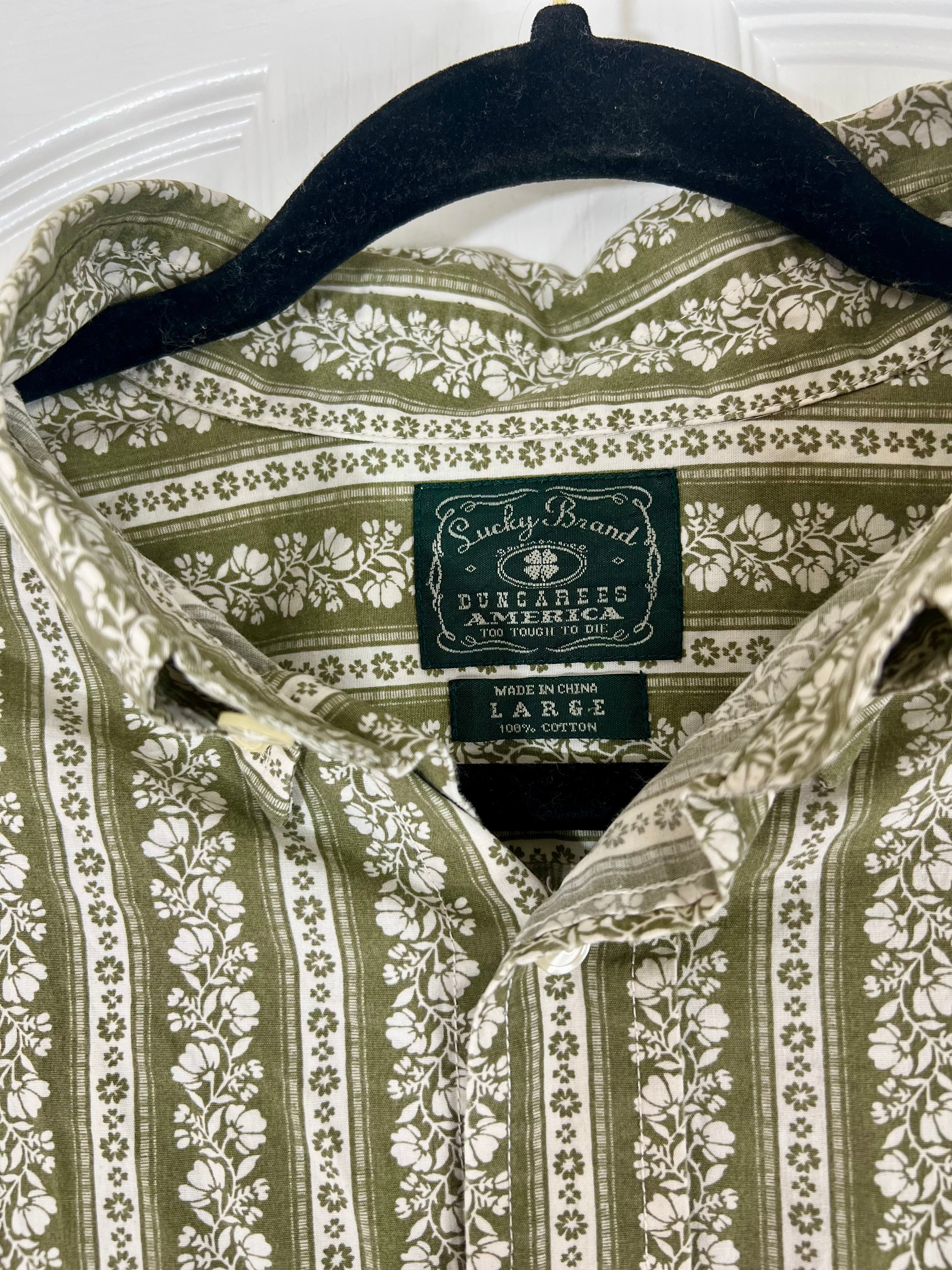 A close-up of a collared button-up shirt hanging on a black hanger, featuring an olive green and cream floral pattern. The neck label clearly shows the brand 'Sucky Brand' and 'Dunlap's America'.