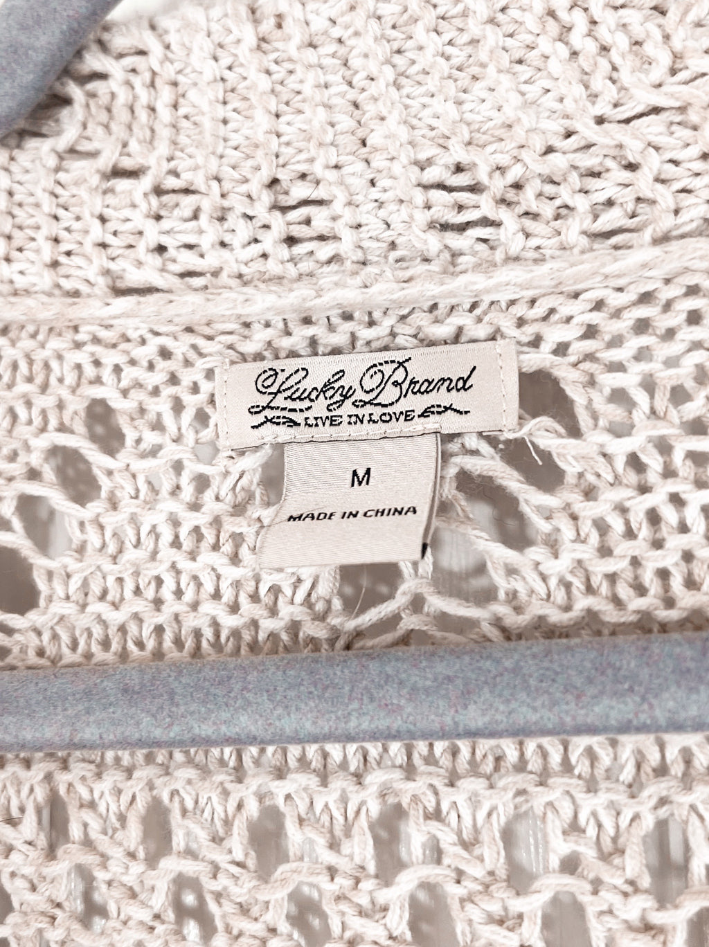 Close-up of a cream-colored crochet knit fabric with a visible sewn-in label reading 'Lucky Brand' and size 'M'.