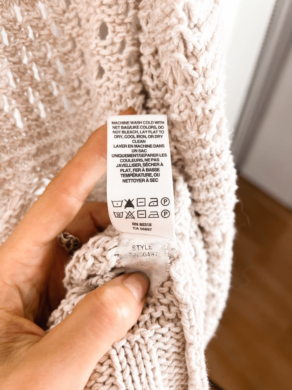 A close-up of a hand holding a white care label attached to a chunky knit sweater in a soft cream or light beige color, with a blurred indoor background.