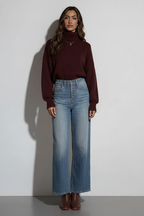 A woman stands against a light gray wall wearing a dark burgundy turtleneck sweater and light blue wide-leg jeans. A 'bitStudio' watermark is visible at the bottom.