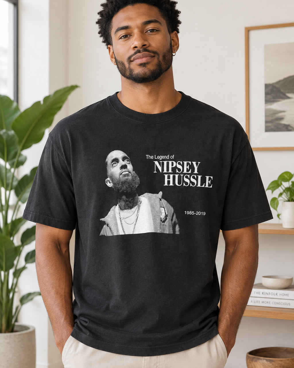 A black short-sleeve graphic t-shirt hanging on a black hanger against a light beige fabric backdrop. The front of the shirt features a black-and-white photo of Nipsey Hussle with white text reading 'The Legend of NIPSEY HUSSELE' and '1965-2019'.