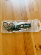 A Los Angeles Lakers keychain inside a clear plastic hanging blister pack, placed on a light-colored wooden table. The keychain is dark green with the Lakers logo and a silver carabiner.