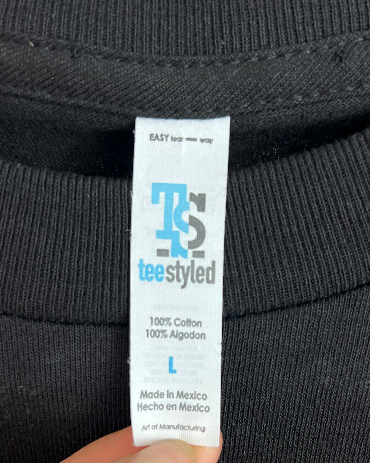 A close-up of a black crewneck sweatshirt showing the inner neck label, held by a hand. The white label features the teal 'teestyled' brand logo and indicates it is made in Mexico.