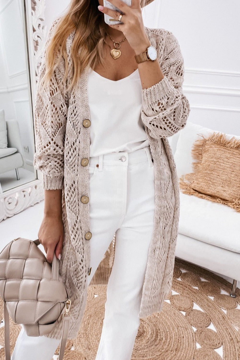 Person wearing a long beige cardigan over a white outfit, holding a beige handbag in a living room.