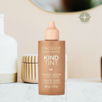 A bottle of Pacifica Kind Tint Tinted Serum in a warm tan shade, displayed on a marble surface with neutral decorative containers in the background.
