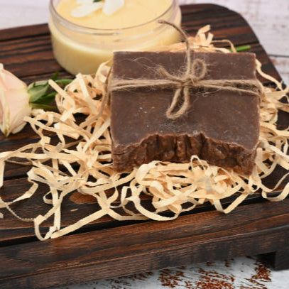 Bar of soap on a wooden board with a small container of cream and a flower.