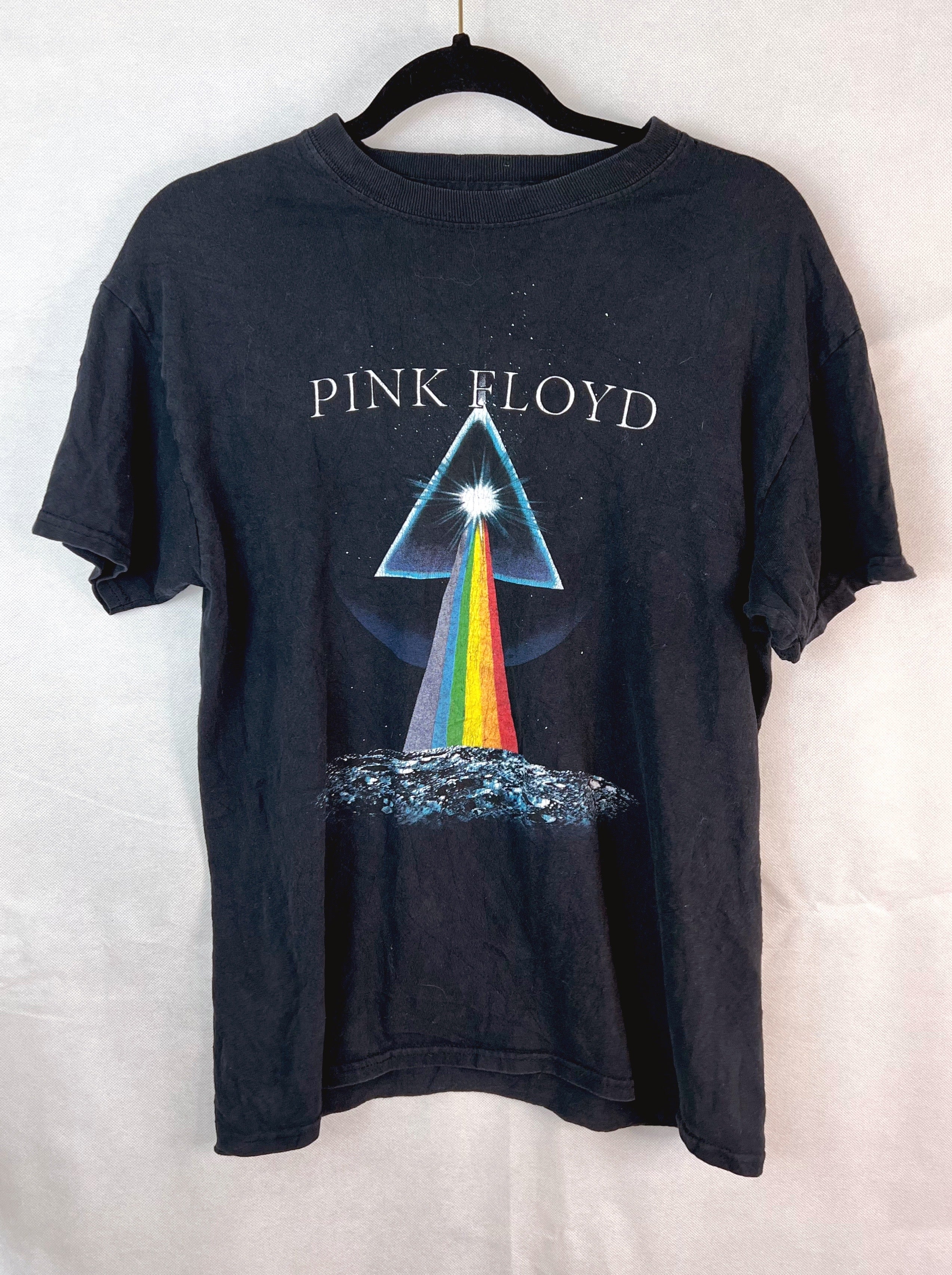 A vintage black Pink Floyd 'The Dark Side of the Moon' graphic t-shirt hanging on a black hanger against a light gray fabric backdrop. The shirt features the band's name in white text above the iconic prism graphic with a rainbow beam of light.