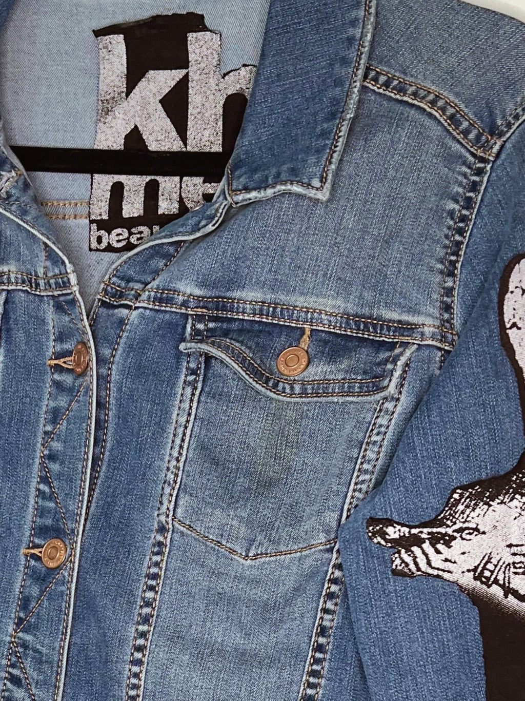 Close-up of a blue denim jacket with a graphic print on the sleeve, showing a pocket, button, and part of a black-and-white wolf design.