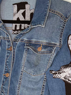 Close-up of a blue denim jacket with a graphic print on the sleeve, showing a pocket, button, and part of a black-and-white wolf design.