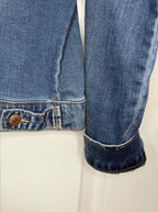Close-up of a blue denim jacket sleeve with a rolled cuff and a buttoned pocket detail, shown against a plain light background.